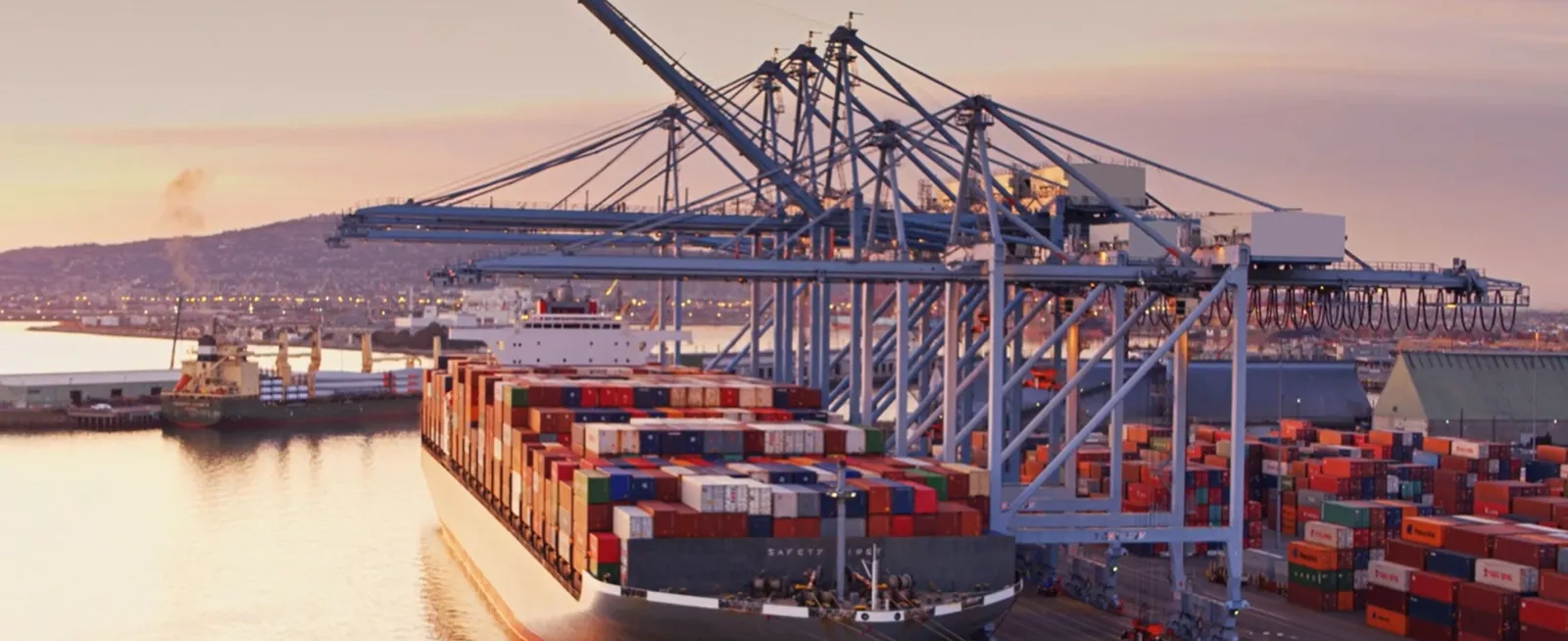 Cargo ship docked at a bustling port with cranes unloading colorful shipping containers. Industrial buildings and a distant hill are visible in the background.